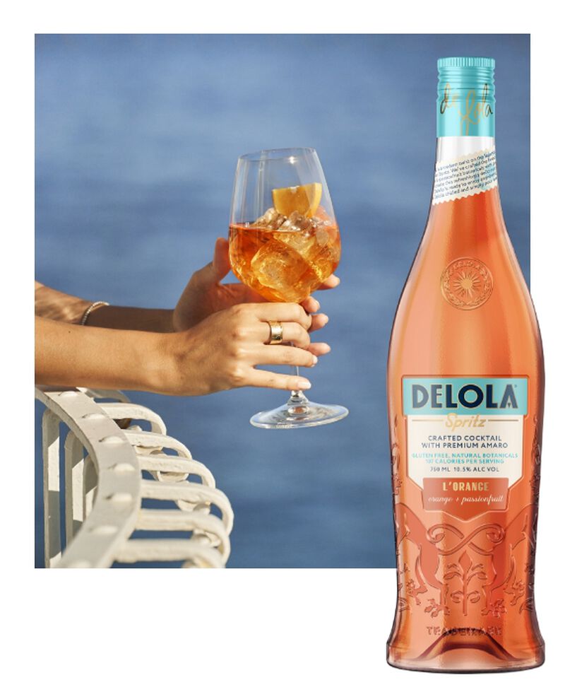 A bottle of Delola L'Orange Spritz with someone enjoying a spritz in a wine glass