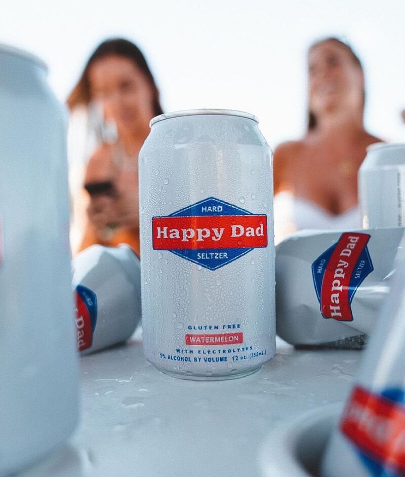 Can of Happy Dad Hard Seltzer with other cans on a table with two friends in the background
