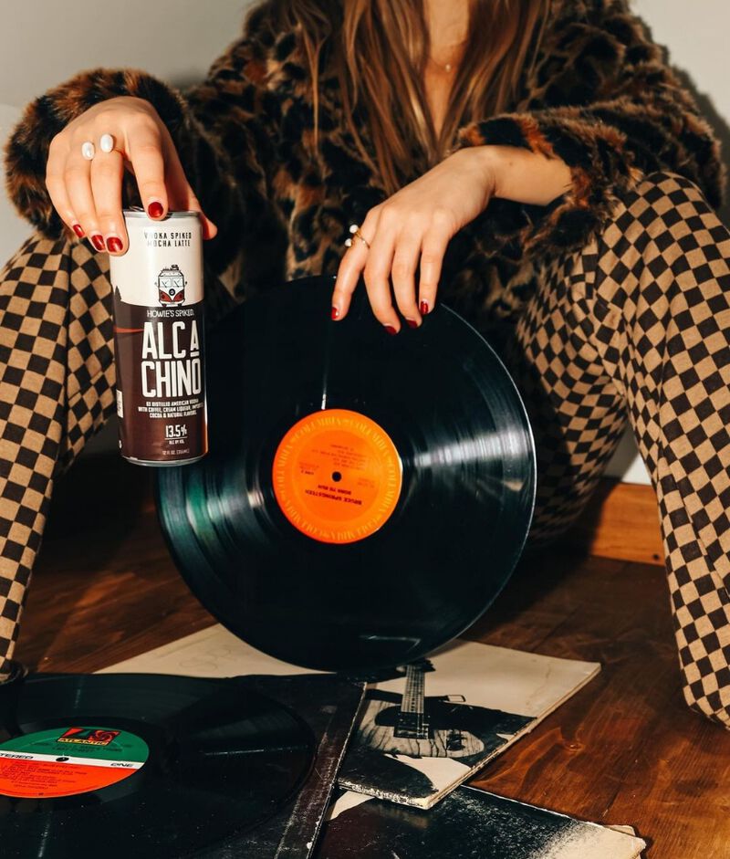 A woman holding a can of  Howie's Spiked Alc-a-Chino Mocha Latte in one hand, and a record in the other
